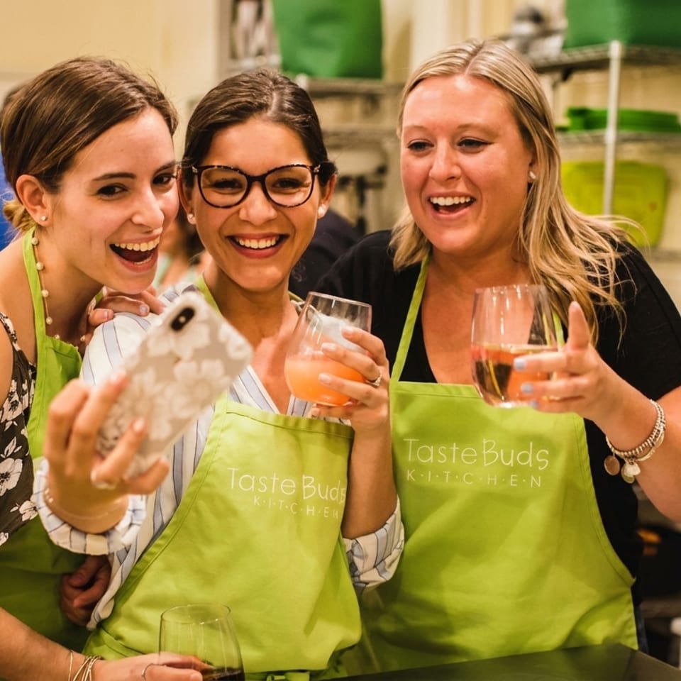 Three smiling women in green Taste Buds Kitchen aprons take a selfie, holding drinks.