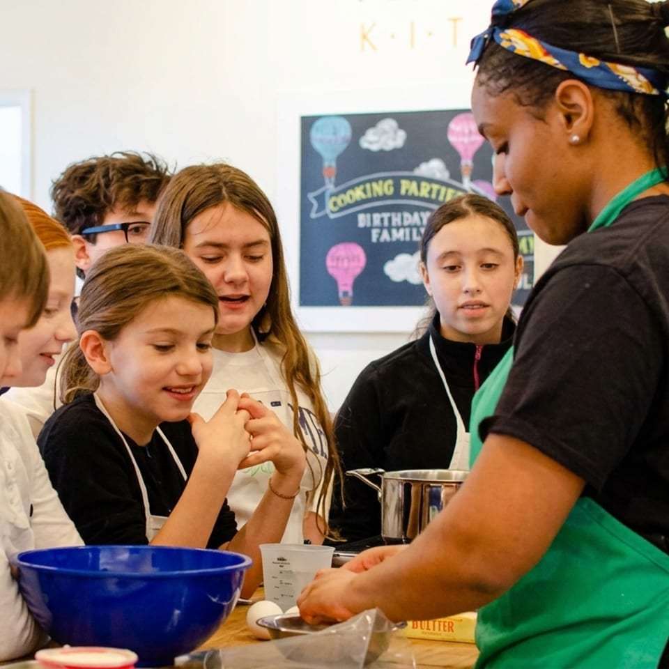 Kids in aprons watch intently as an instructor demonstrates cooking techniques at a class.