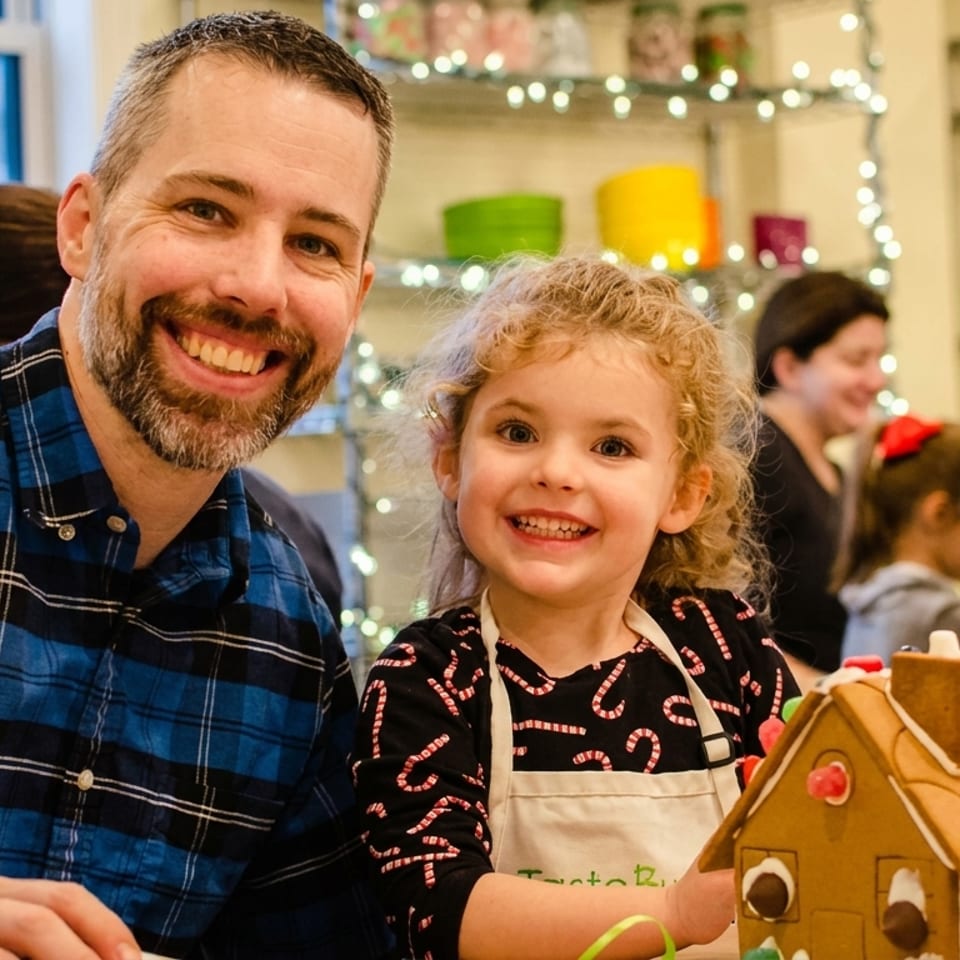 Smiling man and young girl with a decorated gingerbread house at a holiday event.