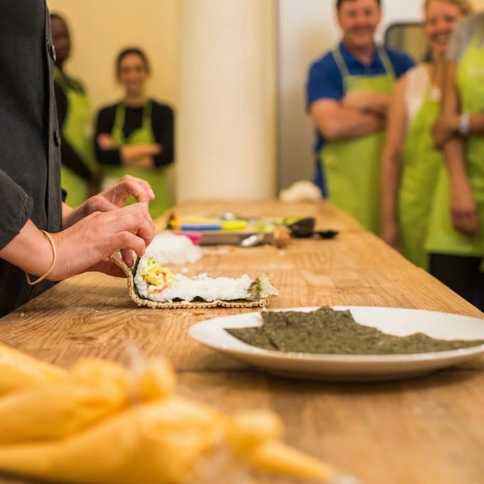 A person making sushi on a wooden table, with other people in green aprons watching.