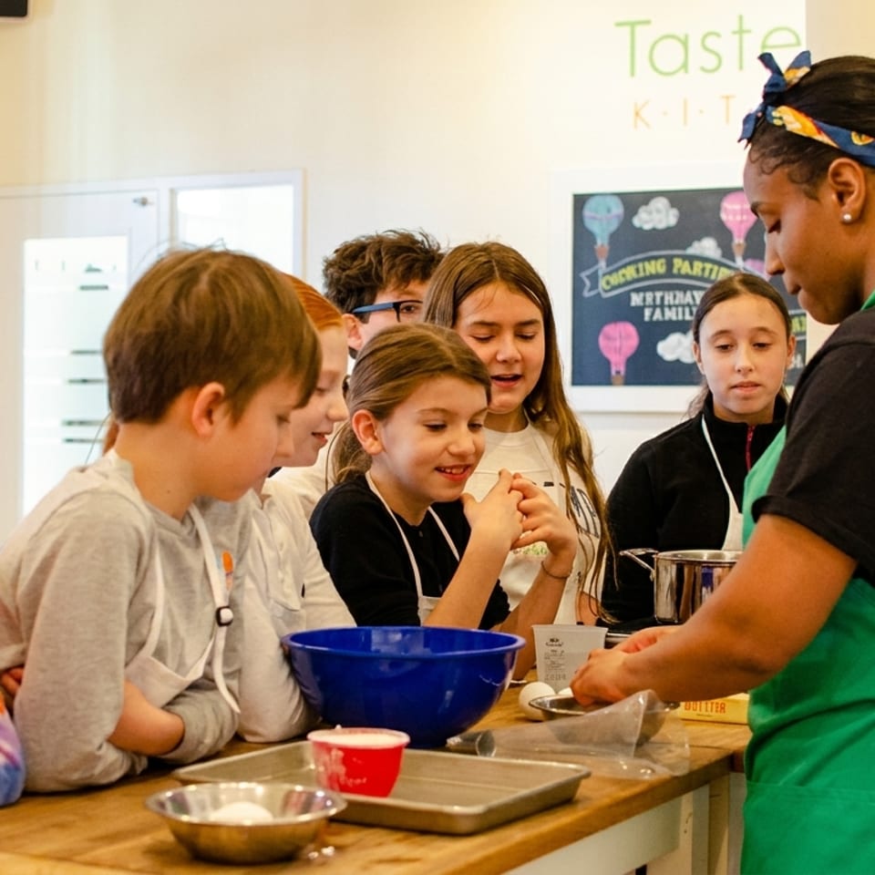 Kids in aprons watch intently as an instructor demonstrates cooking at a kitchen counter.