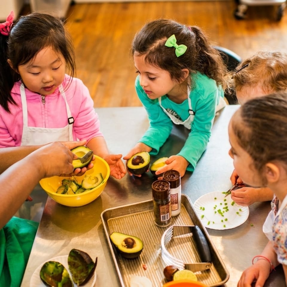An adult and several young children in aprons prepare avocados and other ingredients at a cooking class.