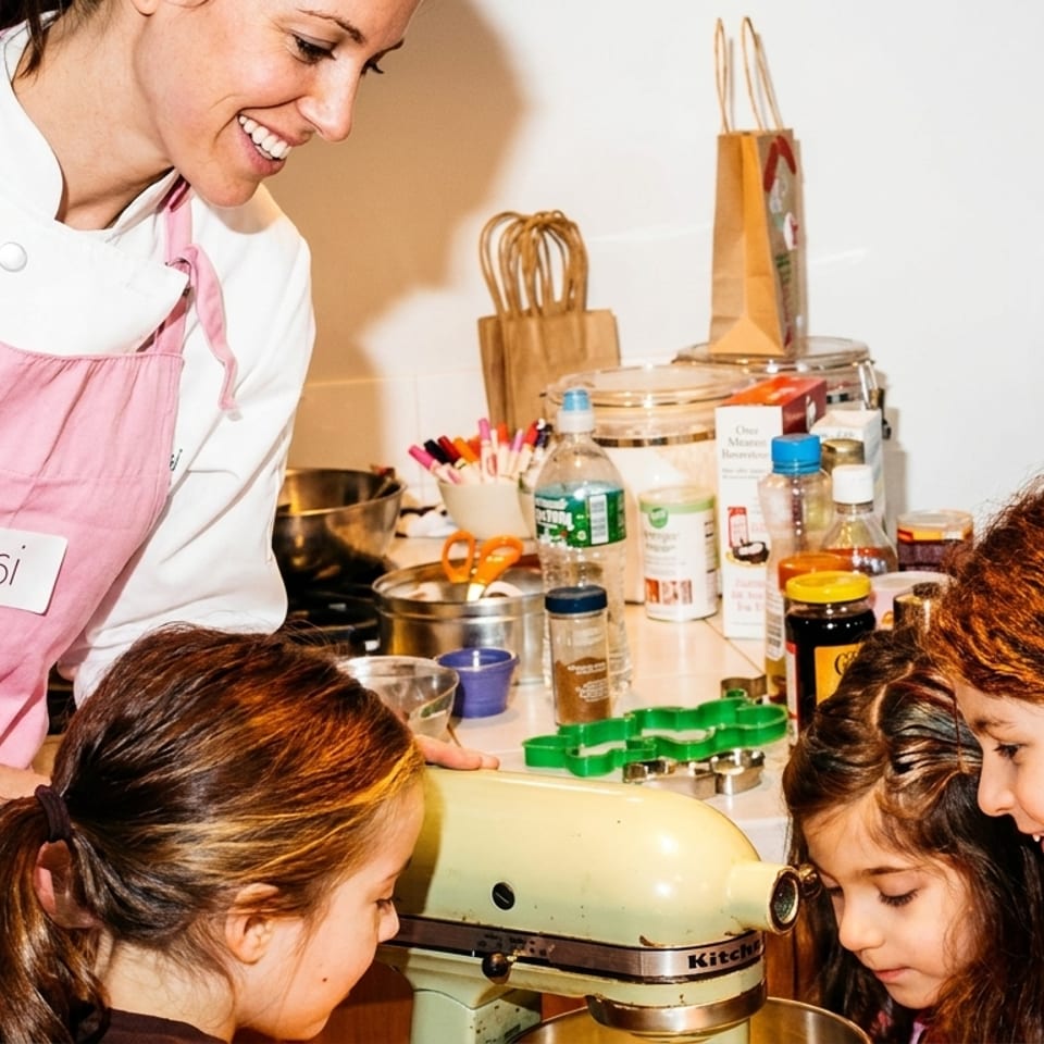 Chef Jessi in a pink apron teaching young girls how to use a KitchenAid mixer.