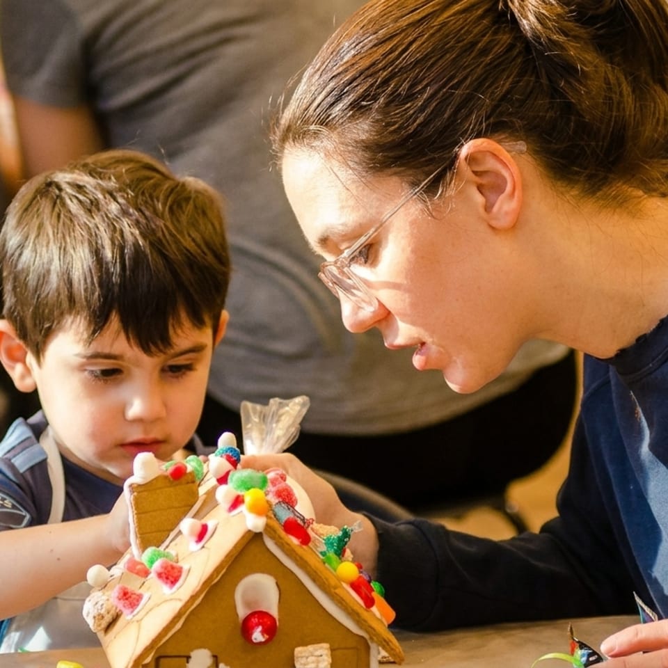 A woman and young boy decorating a gingerbread house with colorful candies and icing.