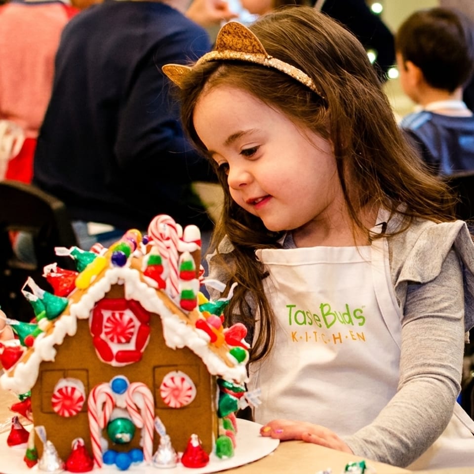 A young girl in a "Taste Buds Kitchen" apron decorates a gingerbread house with candy.
