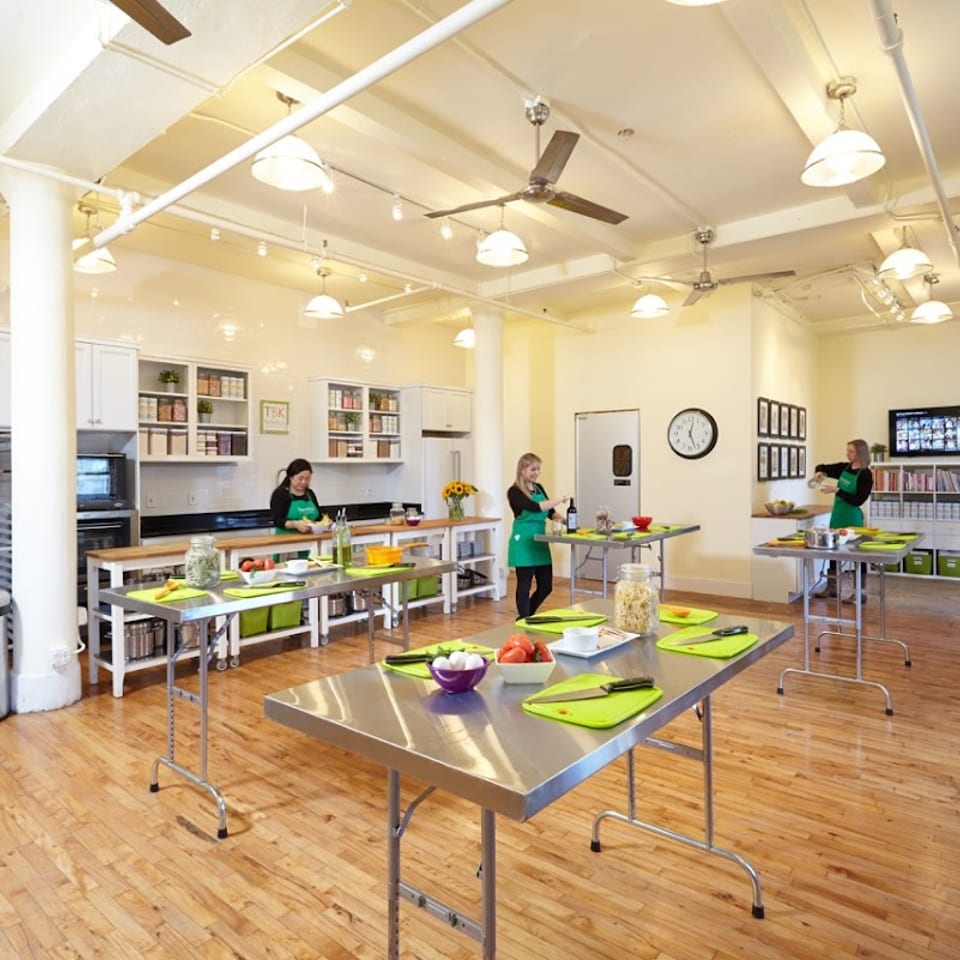 Three women in green aprons prepare food at stainless steel tables in a bright, spacious kitchen.