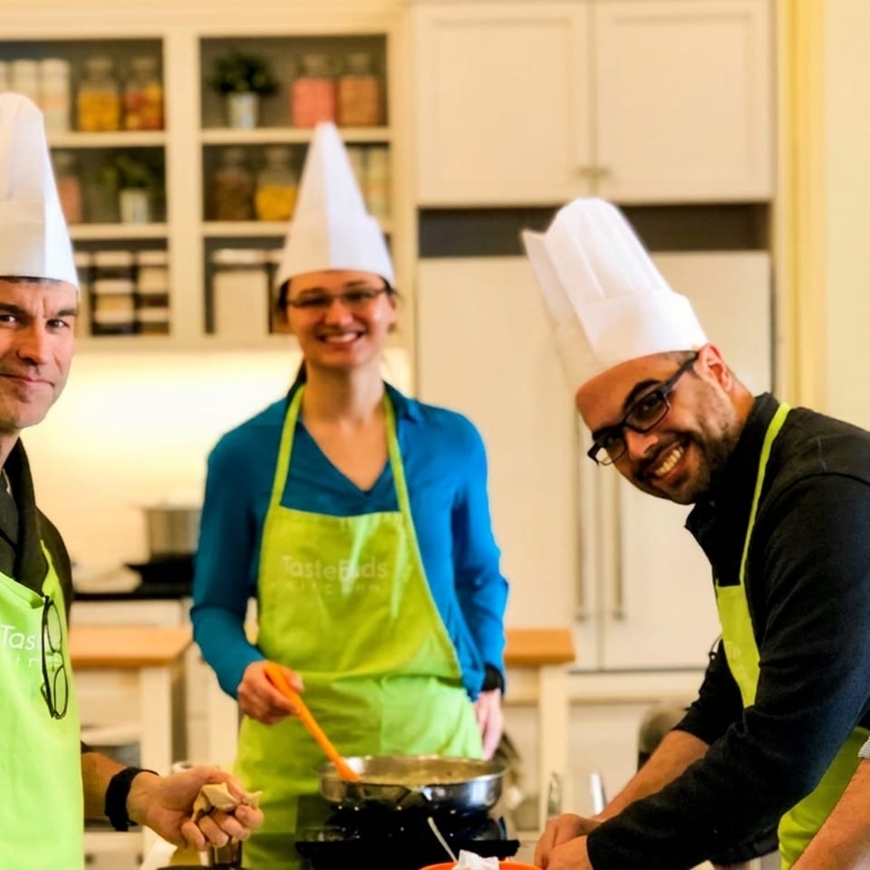 Four smiling adults in chef hats and green aprons cooking together in a bright kitchen.