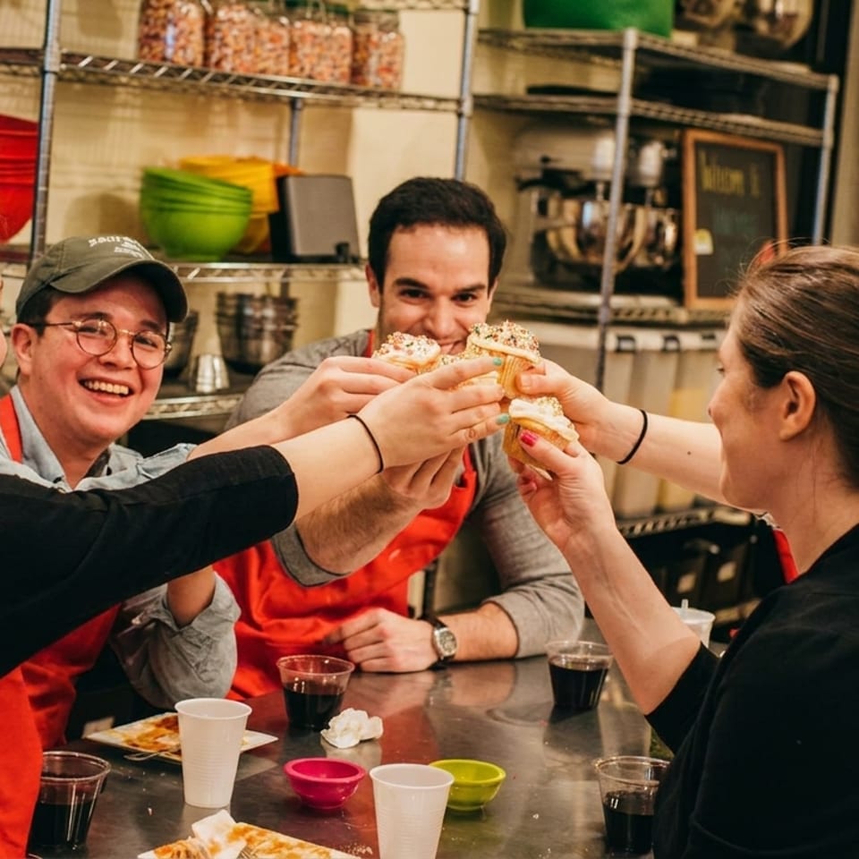 Five people in red aprons cheer with cupcakes around a table in a kitchen.