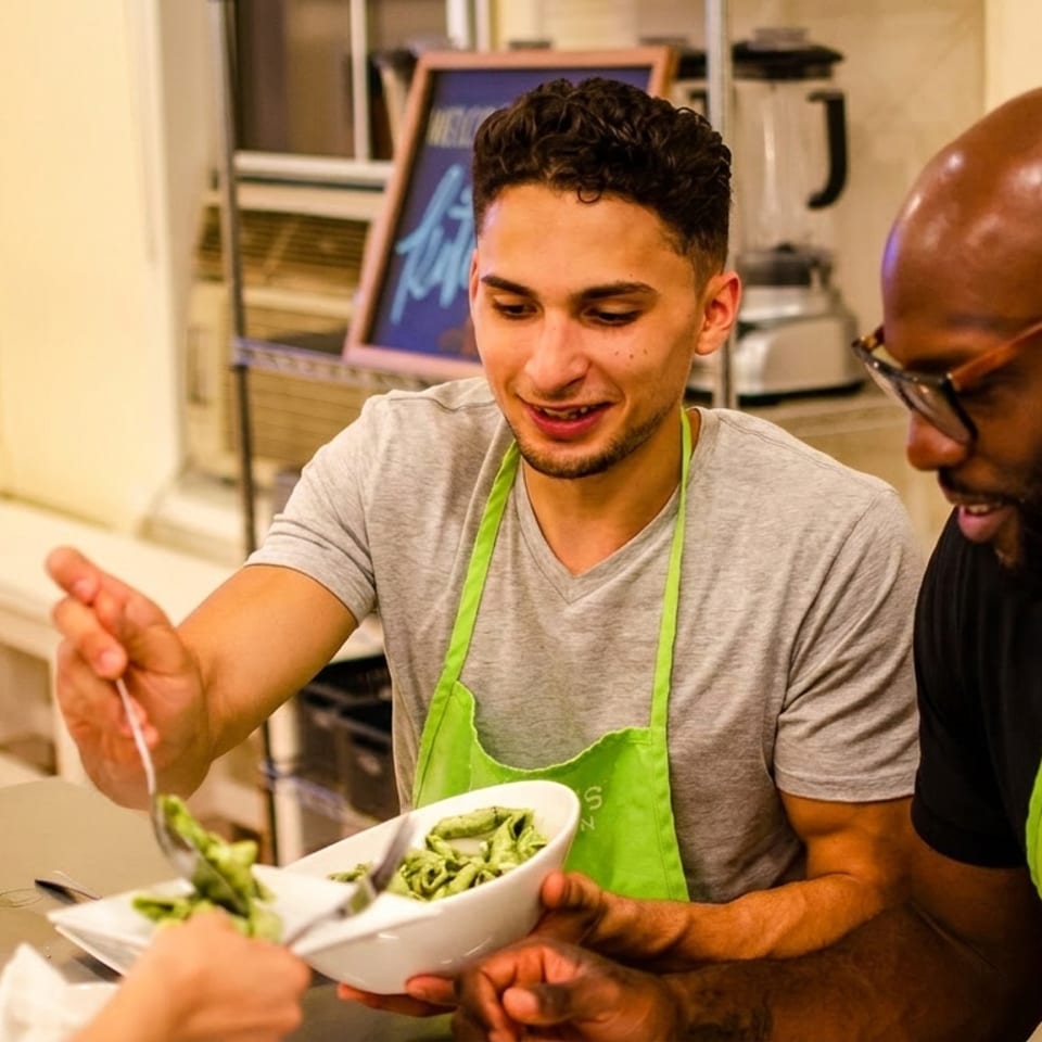 Two men in green aprons serve pasta into a bowl, with wine nearby.