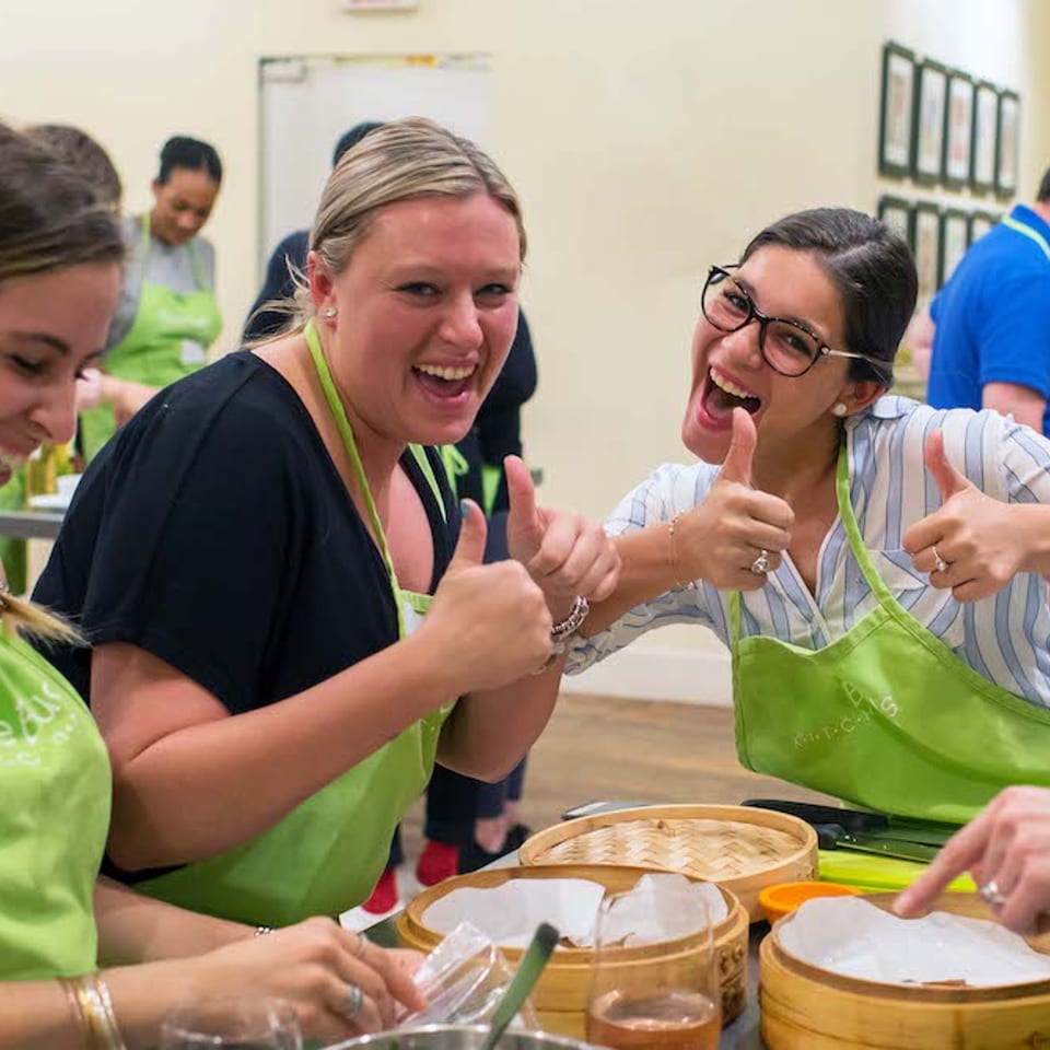 Three women in green aprons laugh and give thumbs-up during a cooking class.