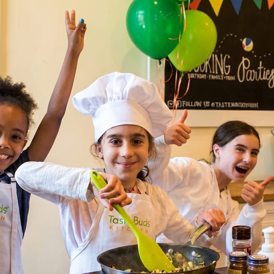 Three smiling girls in aprons and chef hats cooking in a Taste Buds Kitchen class.