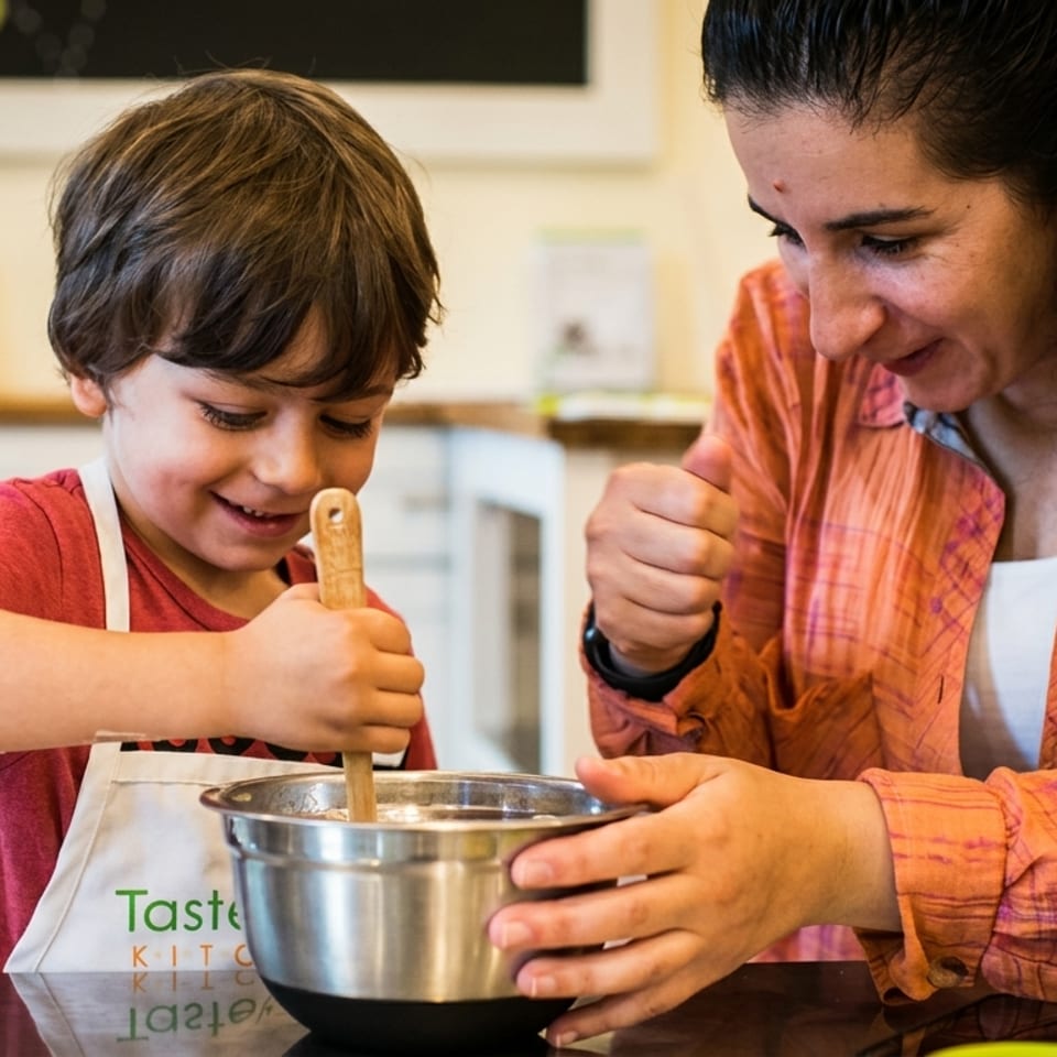 A smiling child in an apron stirs a bowl with a wooden spoon, guided by an adult.